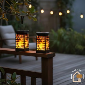 May include: Two black solar-powered lanterns with a flame effect, placed on a wooden railing. The lanterns have a rectangular design with a handle and emit an orange glow. The background features a wooden deck, string lights, and foliage.