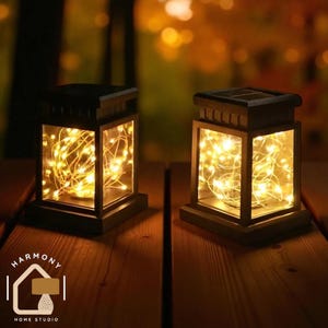 Op de afbeelding: Twee zwarte, rechthoekige zonne-lantaarns met warmwitte lichtslingers. De lantaarns hebben een houten uitstraling en heldere glazen panelen. De lichten zijn zichtbaar door de panelen. Op een houten oppervlak. Het logo "Harmony Home Studio" is zichtbaar.
