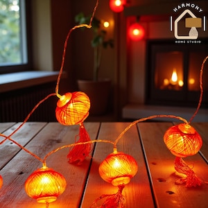 May include: String lights with orange and yellow spherical lanterns, each with a tassel. The lights are on a wooden surface, with a fireplace and plant in the background. The Harmony Home Studio logo is in the upper right corner.