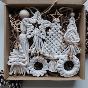 Può includere: Una collezione di ornamenti natalizi in macramè bianco fatti a mano in una scatola di cartone. Gli ornamenti includono stelle, alberi, ghirlande e ali d'angelo, con perline e stelle di legno. La scatola è riempita di carta marrone.
