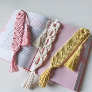 Könnte beinhalten: Drei handgefertigte Makramee-Lesezeichen in Rosa, Creme und Gelb, jedes mit einer Quaste. Die Lesezeichen befinden sich in einem offenen Buch mit rosa Seiten. Die Designs umfassen ein Fischgrätmuster und eine Reihe miteinander verbundener Kreise.
