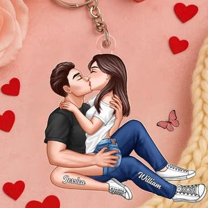 May include: A personalized keychain featuring a couple kissing, with the names "Jessica" and "William" on their legs. The illustration is set against a pink background with red hearts and a butterfly. The couple is wearing casual clothes.