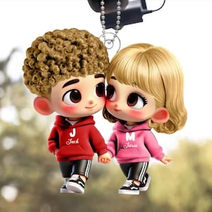 May include: A car charm featuring two cartoon figures, a boy and a girl, holding hands. The boy has curly brown hair and wears a red hoodie with the letter "J" and the name "Jack". The girl has blonde hair and a pink hoodie with the letter "M" and the name "Jaria".