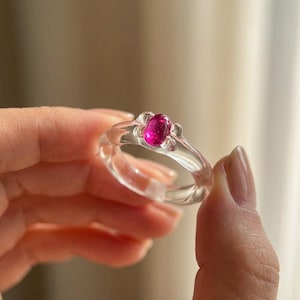 Op de afbeelding: Een heldere ring met een felroze edelsteen in het midden. De ring heeft een delicaat bloemmotief rond de edelsteen. De band is glad en lijkt gemaakt van een transparant materiaal.