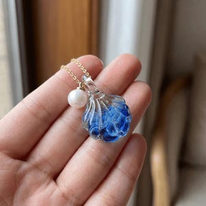 May include: A delicate necklace featuring a pendant shaped like a seashell. The pendant is clear with blue accents, and a small white pearl hangs next to it. The necklace has a thin gold chain.
