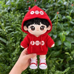 May include: A plush doll with black hair, wearing a red hooded sweatshirt featuring white circular and horizontal line graphics on the chest and hood. The doll has large brown eyes, rosy cheeks, and wears white and red low-top canvas sneakers.