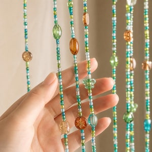 Pode incluir: Close-up de fios de contas coloridas em tons de verde, azul e âmbar. As contas são de várias formas e tamanhos, com desenhos ovais e florais. Os fios são amarrados, criando um elemento decorativo.