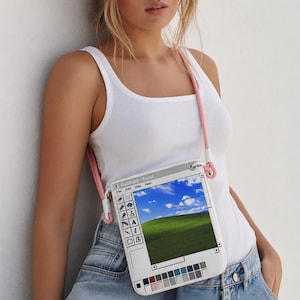 Könnte beinhalten: Eine rechteckige weiße Umhängetasche mit einem Grafikdruck einer klassischen Computer-Malsoftware-Oberfläche. Das Design zeigt eine Landschaft mit blauem Himmel und grünen Hügeln, eine Werkzeugleiste mit verschiedenen Symbolen und einen rosa Seil-Schulterriemen.