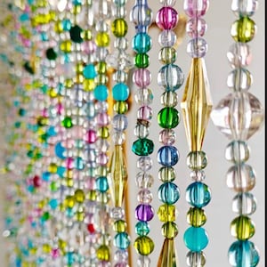 May include: A beaded curtain with strands of colorful beads in shades of pink, blue, green, yellow, and clear. Some strands have gold-colored diamond-shaped beads. The beads are round and of varying sizes, creating a decorative and eye-catching design.
