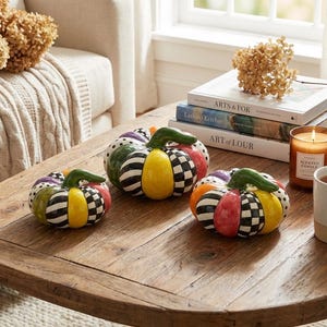 Pode incluir: Três abóboras decorativas de cerâmica com padrões coloridos, incluindo listras, xadrez e bolinhas, estão em uma mesa de centro de madeira. Cada abóbora tem um caule verde. Uma vela, livros e uma caneca também estão na mesa.