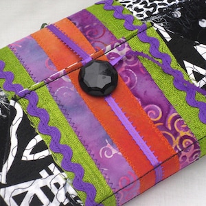 May include: A colorful fabric clutch with a black button closure. The clutch is made of different fabrics, including purple, orange, green, and black. The fabric is sewn together with a zig-zag stitch.