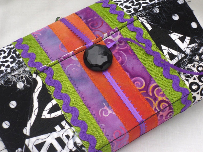May include: A colorful fabric clutch with a black button closure. The clutch is made of different fabrics, including purple, orange, green, and black. The fabric is sewn together with a zig-zag stitch.