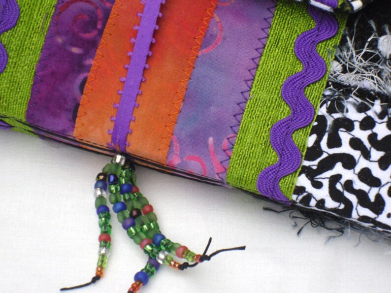May include: A purple, orange, and green fabric patchwork with a beaded tassel. The fabric is sewn together with a zig-zag stitch.