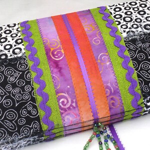 May include: A colorful patchwork fabric pouch with a purple, green, and orange ribbon trim. The pouch has a black and white patterned fabric with a fringe edge. A beaded tassel hangs from the bottom of the pouch.
