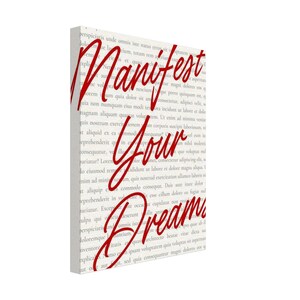 Peut inclure: Une toile murale rectangulaire présentant un fond beige clair recouvert de petits textes latins gris répétitifs. Au centre, la phrase "Manifest Your Dreams" est inscrite en lettres cursives rouges, grasses et bien visibles.