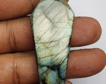 May include: A coffin-shaped labradorite gemstone with a polished surface. The stone displays a mix of colors, including blue, green, and gray, with a shimmering effect. The gemstone is held in a hand against a white background.
