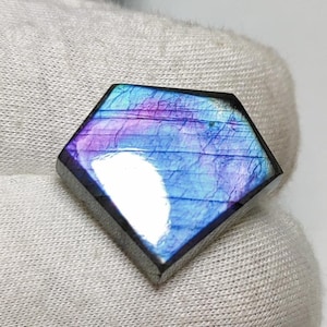May include: A diamond-shaped gemstone with a shimmering display of colour. The stone features blues, purples, and greens. It is set in a silver-coloured metal. The background is a white surface.