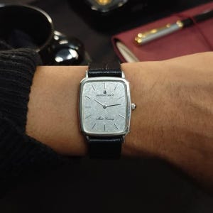 May include: A silver-toned Universal Geneve Mark Century wristwatch with a rectangular case, a textured silver dial, and a black leather strap. The watch face features thin black hour markers, black hands, and the text Universal Geneve Mark Century.