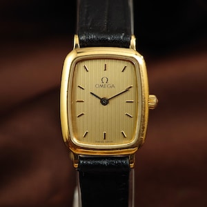 May include: A gold-tone rectangular wristwatch with a textured gold dial, black hour markers, and black hands. The watch features the Omega logo and text at the top, with Swiss Made printed at the bottom. It is attached to a black leather strap.