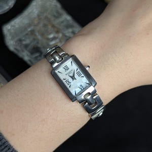 Puede incluir: Un reloj de pulsera rectangular plateado con esfera blanca y números romanos. El reloj presenta una esfera de nácar y está marcado con "Balmain Swiss". Se sujeta con una pulsera de eslabones plateada.