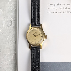 May include: A vintage gold-toned Omega Genève wristwatch with a black leather band. The watch face is a light gold color with black hands and markers. The watch has the Omega logo and the word "Genève" on the face.