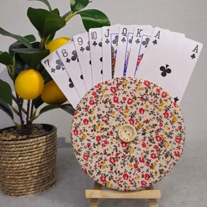 Puede incluir: Un tarjetero decorativo con un diseño de tela floral, que sostiene una mano de naipes. El soporte está sobre un pequeño caballete de madera. Un pequeño limonero en maceta está en el fondo.