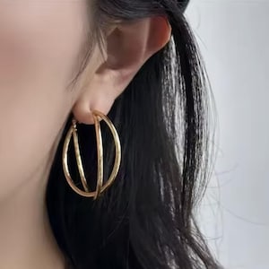 Könnte beinhalten: Goldfarbene Creolen mit einem einzigartigen dreidimensionalen Design. Die Ohrringe haben einen kreisförmigen Reifen, der von zwei geraden Stäben gekreuzt wird, wodurch ein moderner und stilvoller Look entsteht. Der Schmuck wird von einer Person getragen.