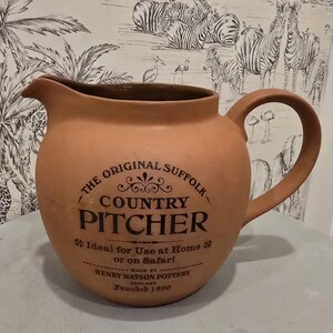 May include: A terracotta pitcher with a curved handle and a spout. The front features black text reading THE ORIGINAL SUFFOLK COUNTRY PITCHER, Ideal for Use at Home or on Safari, MADE BY HENRY WATSON POTTERY ENGLAND, and Founded 1800.