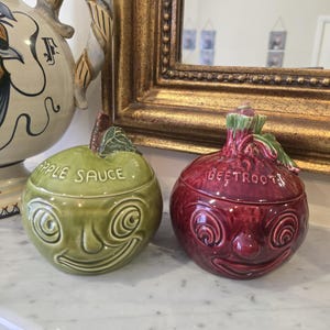 May include: Two ceramic lidded jars shaped like vegetables with faces. The green jar is labelled APPLE SAUCE and the deep red jar is labelled BEETROOT. Both feature sculpted leaves on the lids and embossed facial features with swirling eyes.