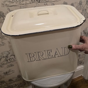 May include: A cream-coloured rectangular metal bread bin with a matching lid and a handle on top. The word BREAD is printed in black capital letters on the front. The edges of the container and lid feature a dark, worn black trim finish.