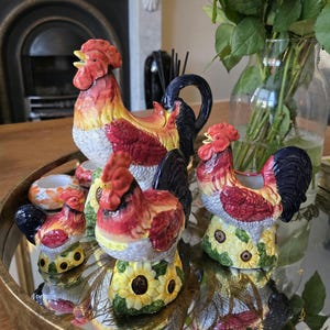 May include: A collection of four ceramic cockerel figurines in varying sizes, featuring red, yellow, and white plumage with dark blue tails. Each cockerel is perched on a base decorated with yellow sunflowers and green leaves.