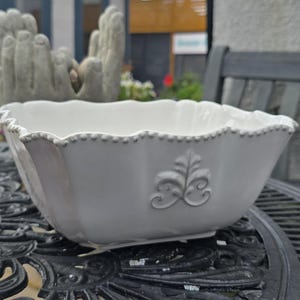 May include: A white ceramic bowl with a scalloped edge and embossed floral pattern. The bowl has a square shape with gently curved sides and a decorative design. The bowl is suitable for serving or display.
