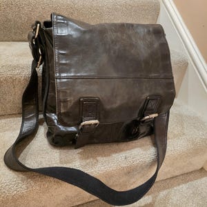 May include: A dark brown leather messenger bag featuring a large front flap, two silver-toned metal buckles, and an adjustable black fabric shoulder strap. The leather surface displays a natural, worn texture with visible creases and subtle variations.