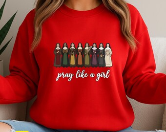 May include: Red sweatshirt featuring a line of illustrated figures in various colored dresses. The phrase "pray like a girl" is written in white script below the figures. The word "RED" is in a yellow box in the lower left corner.