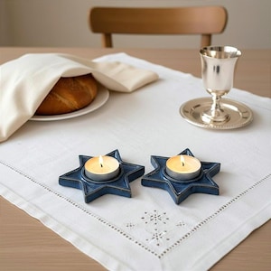 Puede incluir: Una mesa puesta con un mantel blanco, con dos candelabros azules en forma de estrella con velas encendidas, una copa de plata, un plato con pan y una servilleta blanca.