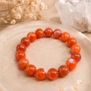 Puede incluir: Una pulsera elástica circular compuesta por cuentas esféricas pulidas en varios tonos de naranja, rojo y blanco translúcido. Las cuentas presentan patrones de bandas naturales y un acabado suave y brillante, dispuestas en un bucle continuo.