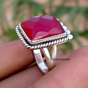 May include: A ring featuring a large, square, faceted deep red gemstone set in a silver frame with a twisted rope border. The band is made of two split silver shanks that meet at the base. The text DevJewelsIndia is visible on the metal.
