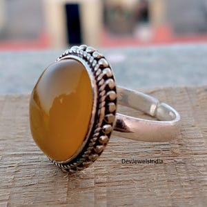 May include: A silver ring featuring an oval, amber-coloured gemstone. The gemstone is set in a decorative bezel with a beaded edge. The ring band is adjustable. The ring is displayed on a wooden surface.