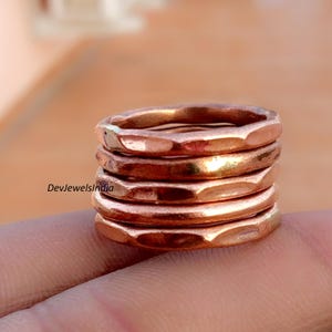 Puede incluir: Una pila de cinco anillos de cobre con un acabado martillado y texturizado. Cada anillo presenta un tono metálico marrón rojizo y una forma redondeada ligeramente irregular. El texto DevJewelsIndia es visible en el lateral.