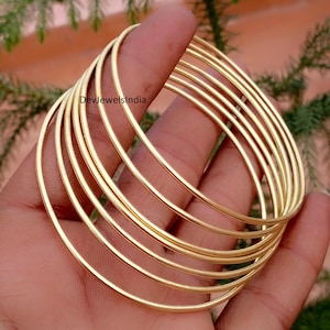 Puede incluir: Un conjunto de siete brazaletes finos, lisos y pulidos de metal en tono dorado apilados juntos. Los brazaletes tienen un diseño simple, redondo y similar a un alambre con una superficie reflectante. El texto DevJewelsIndia es visible en uno.
