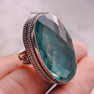 Pode incluir: Um anel oval grande com uma pedra preciosa facetada em tom azul-esverdeado, montado numa banda de prata decorativa com padrões metálicos intrincados nas bordas. O engaste inclui uma borda de contas em torno da pedra translúcida.