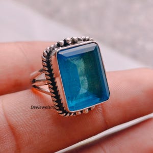 May include: A silver ring featuring a large, rectangular, faceted blue gemstone set in a frame with a twisted rope-like border. The metal band has a split design, and the text DevJewelsIndia is printed in small black letters on the side.