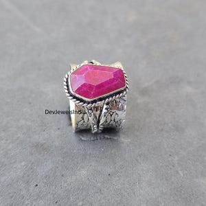May include: A silver ring with a large, faceted, magenta gemstone. The gemstone is rectangular with angled edges, set in a twisted rope-like bezel. The band has a textured, floral design. The text "DevjewelsIndia" is visible on the ring.
