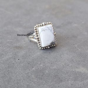 May include: A rectangular silver ring featuring a white stone with gray veining. The stone is framed by a beaded border. The ring is set on a gray surface. The text "DevJewelsIndia" is visible on the ring.