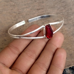 Może przedstawiać: Srebrna bransoletka typu cuff z żywym, prostokątnym, głęboko czerwonym kamieniem szlachetnym pośrodku. Bransoletka ma podzielony design, z kamieniem szlachetnym osadzonym między dwoma równoległymi pasmami. Widoczny jest tekst "DevjewelsIndia".