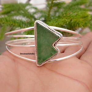 Peut inclure: Un bracelet manchette en argent avec une pierre verte triangulaire au centre, entourée d'une bordure en métal torsadé. Le bracelet est composé de trois fines bandes d'argent parallèles reliées à la pierre centrale. Le texte DevJewelsIndia est visible.