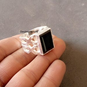 May include: A silver ring with a large, square black stone. The ring has a geometric design on the band. The ring is shown on a hand. The text "DevjewelsIndia" is visible on the ring.