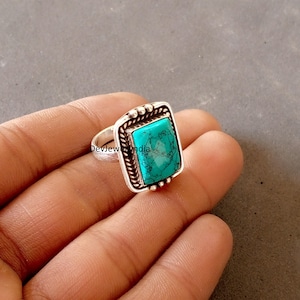 May include: A silver ring with a square, turquoise stone. The ring has a decorative border and is held in a hand. The turquoise stone is a vibrant teal color. The ring is a piece of jewelry.