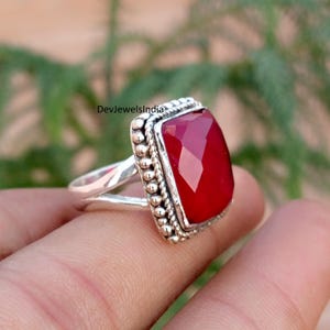 May include: A silver ring featuring a large, rectangular, faceted red gemstone set in a frame with a beaded border. The band is split into two sections where it meets the setting. The text DevJewelsIndia is visible on the metal surface.