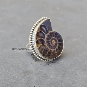 May include: A silver ring featuring a polished ammonite fossil. The fossil displays a spiral pattern in shades of brown and tan, set within a beaded silver band. The ring is a unique piece of jewelry.